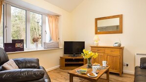A cozy living room with a leather sofa, a wooden coffee table with books and a cup, a TV on a stand, and a window with curtains. - 2 Riverside Cottage (Ambleside)