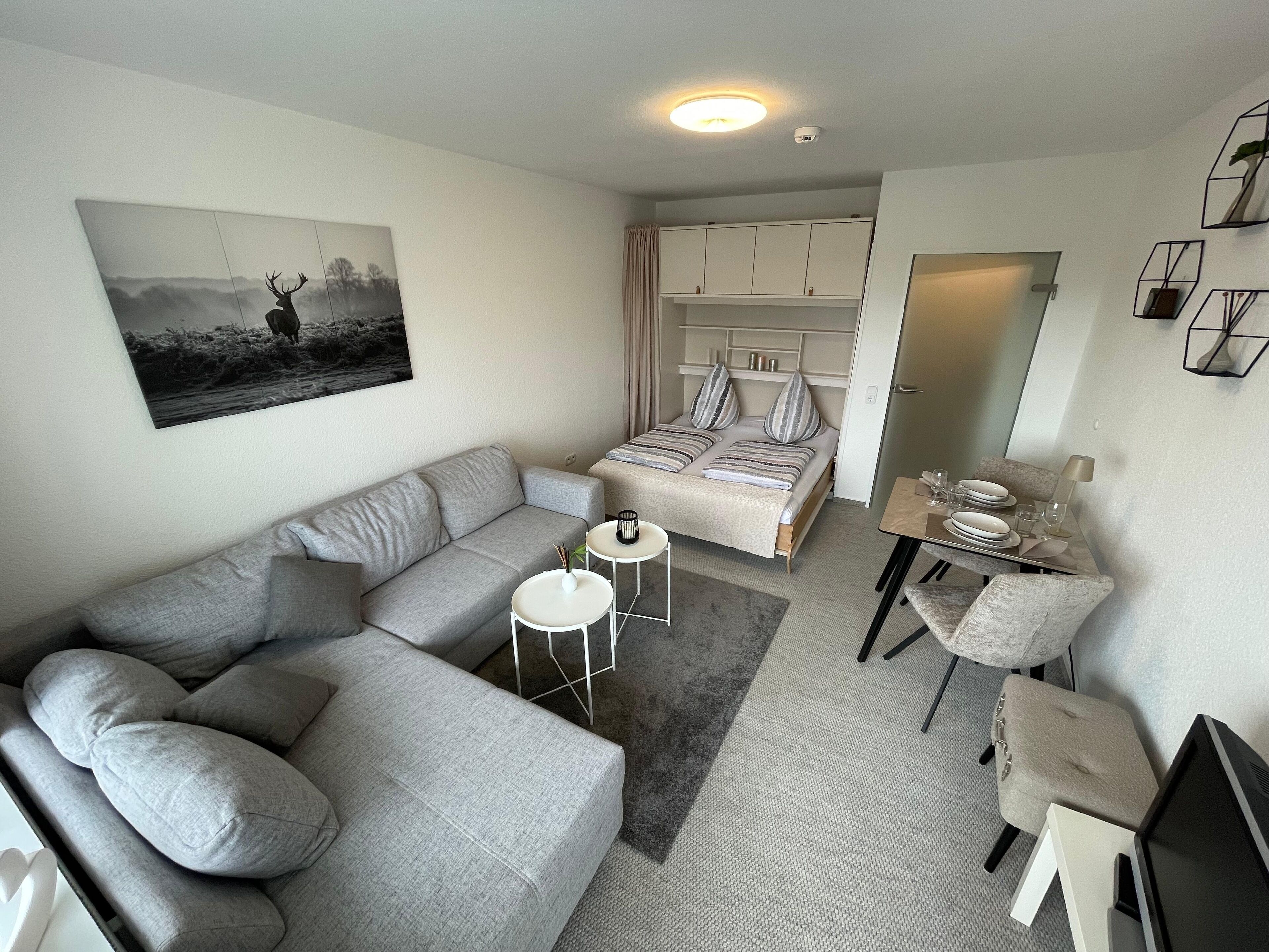 A modern, compact living space with a grey sofa, a small round table, a bed with striped bedding, and a dining area with a table set for two.