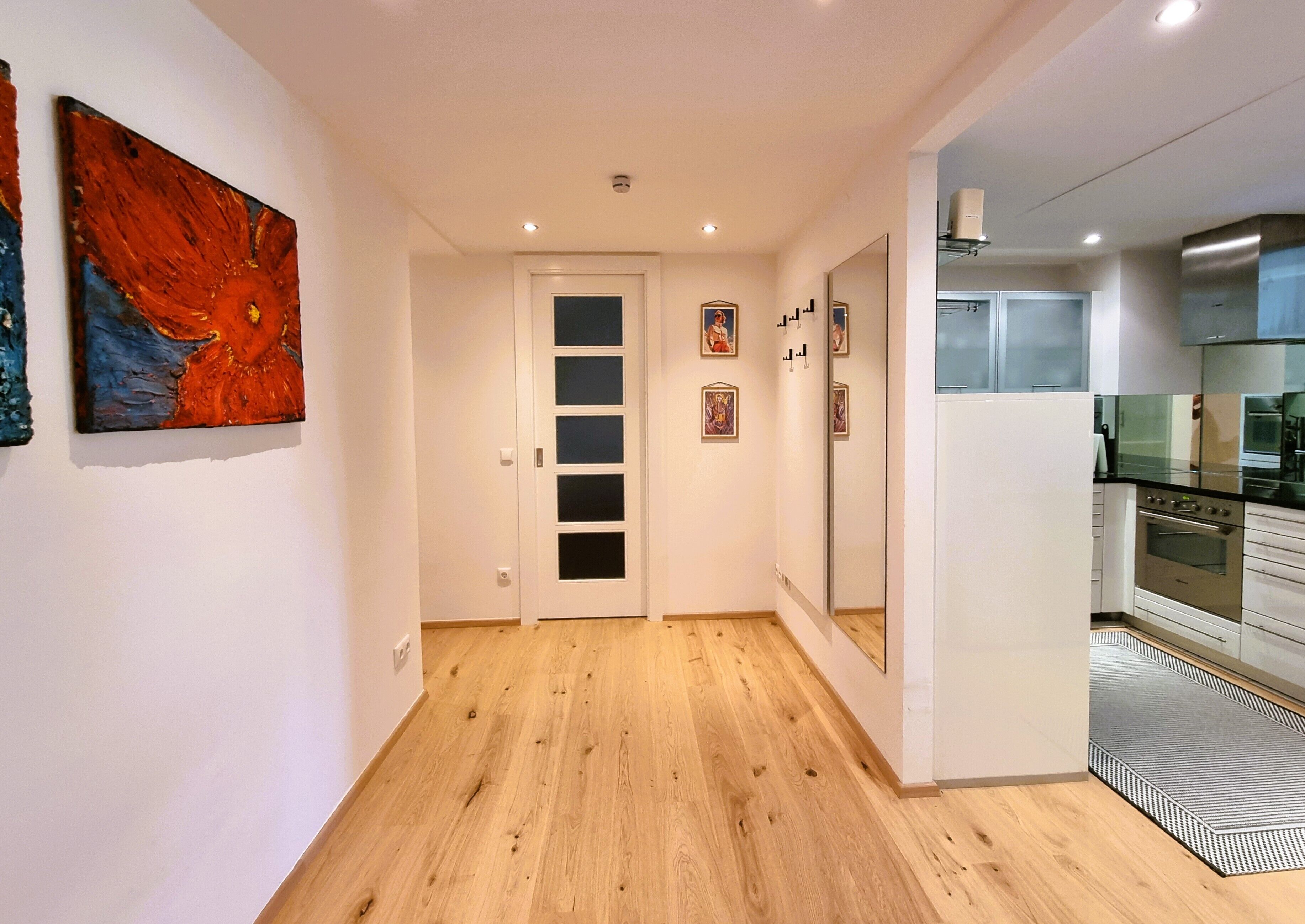 A modern apartment hallway with wooden flooring, a white door, and a kitchen with stainless steel appliances.