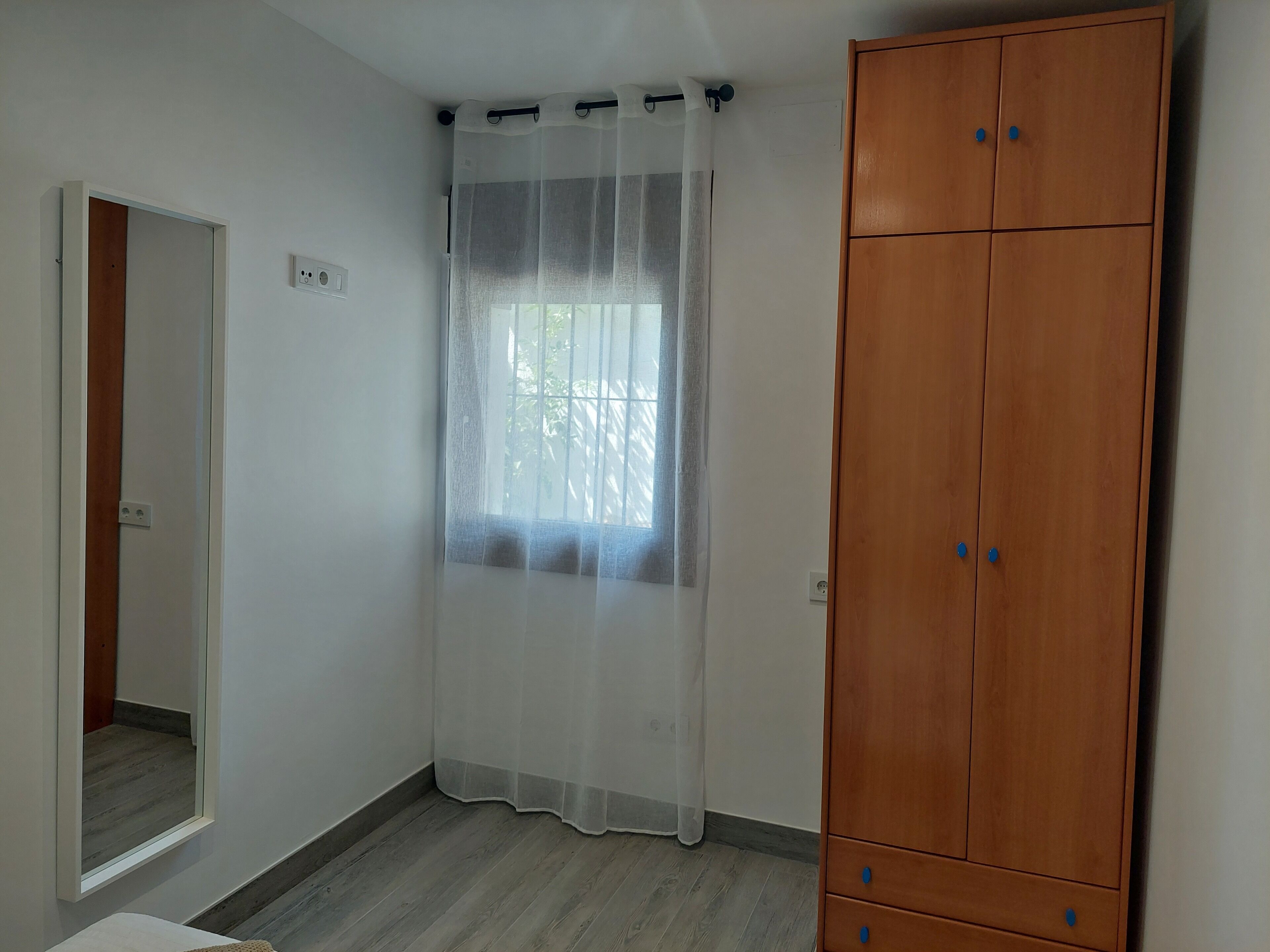 Una habitación con un armario de madera, una ventana con cortinas translúcidas y una puerta.