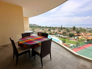 Un balcon avec une table ronde et des chaises, donnant sur un quartier résidentiel avec des maisons et un terrain de sport.