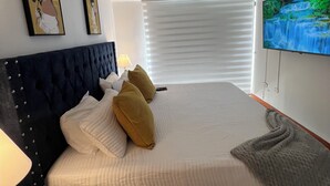 A neatly made bed with a white comforter and two pillows, a dark headboard, a wall-mounted TV, and a window with blinds. - Spacious 4-Bedroom House with AC, Double Parking & Coffee Region Charm (Pereira)