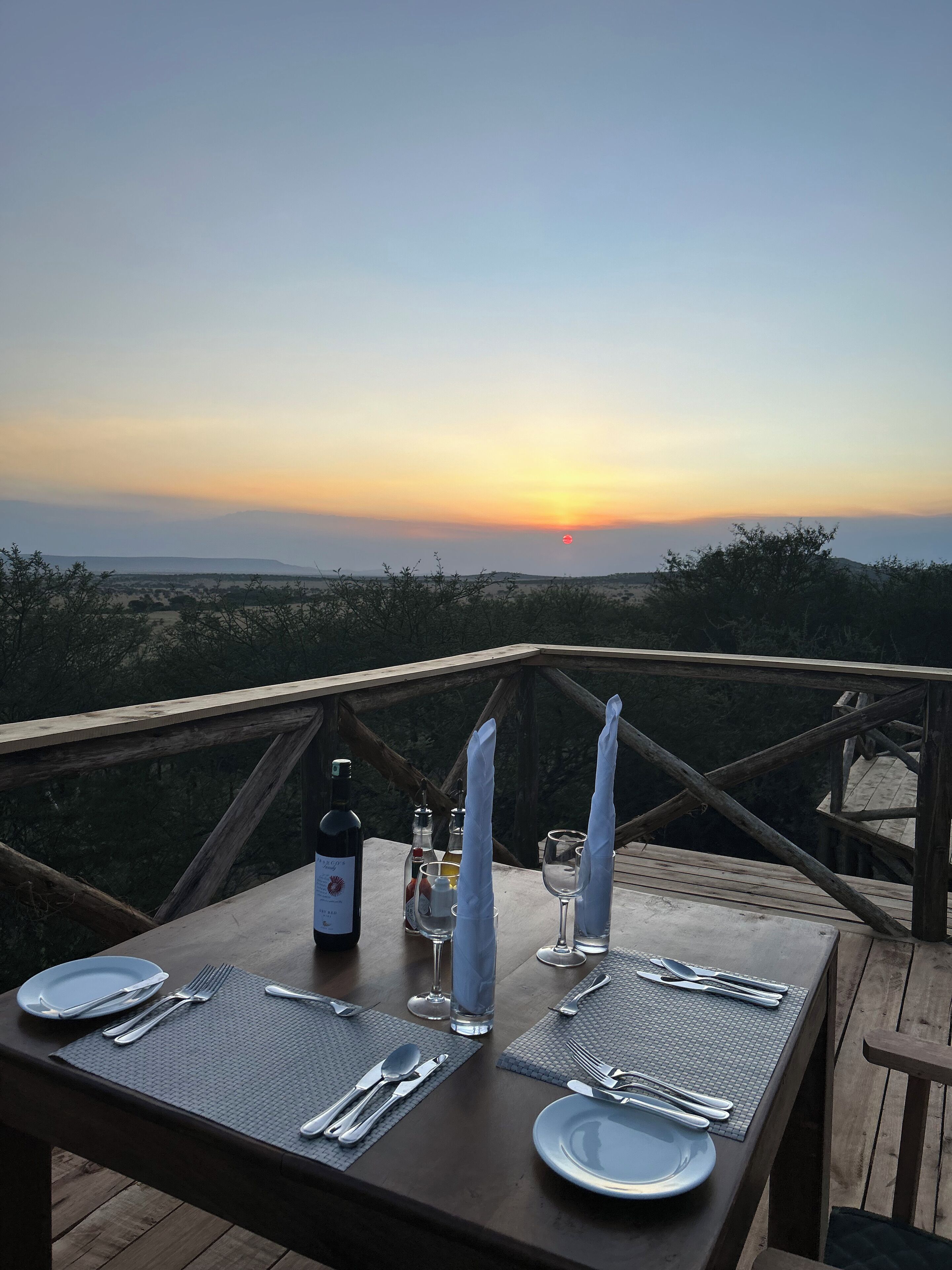 A table set for two with a bottle of wine, glasses, and cutlery, overlooking a scenic sunset view.