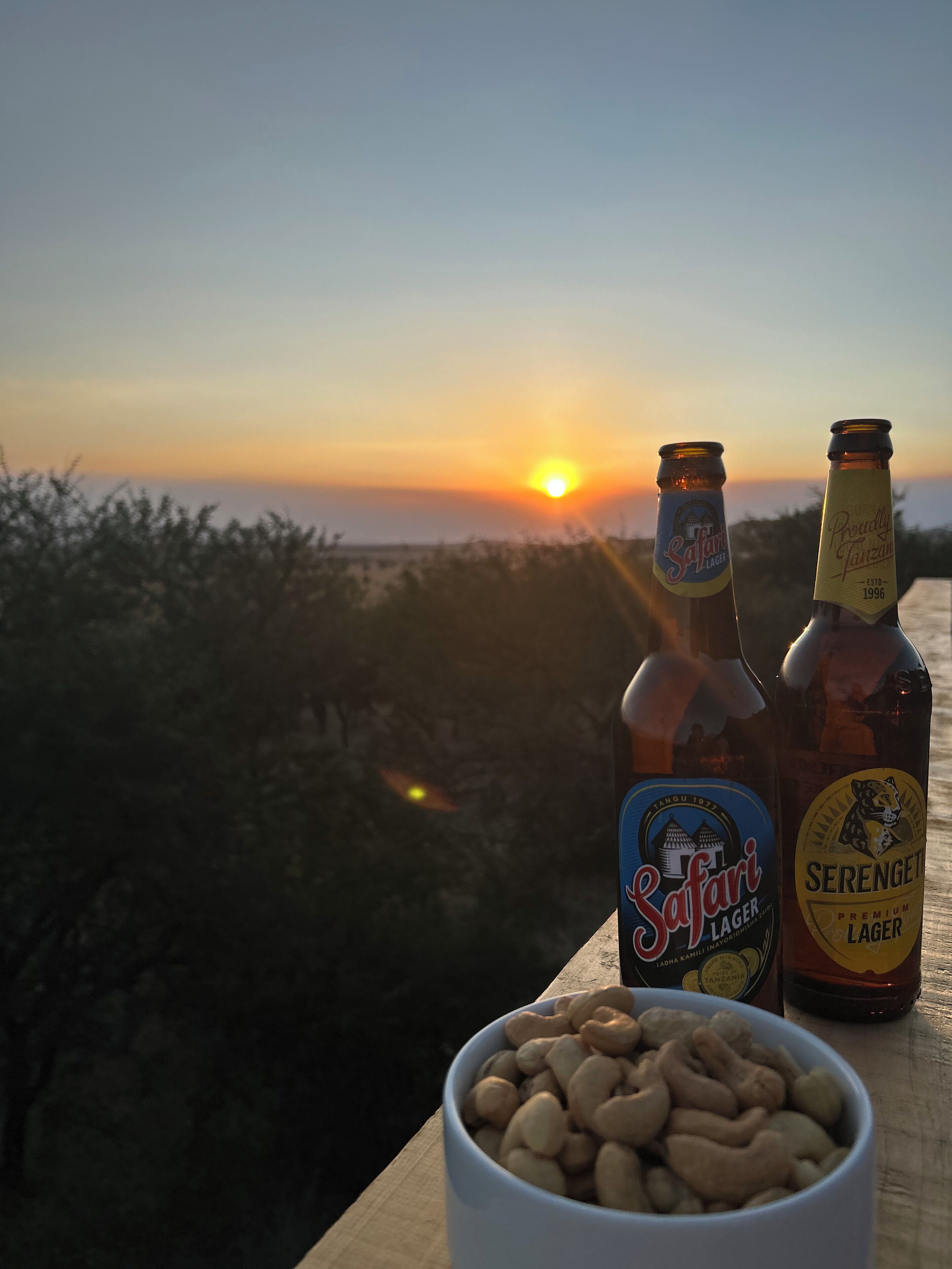 Dos botellas de cerveza y un bol de cacahuates sobre una superficie de madera, con un atardecer de fondo.