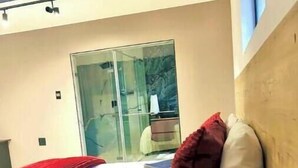 Un dormitorio moderno con una cama, una mesita de noche de madera, un espejo y una puerta de cristal que da al baño, y una almohada roja sobre la cama.