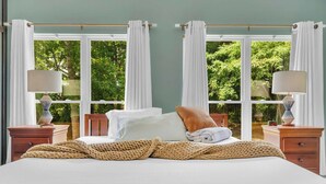 A bedroom with a bed, two nightstands, a window with curtains, and a view of greenery outside.