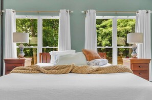 Un dormitorio con una cama, dos mesitas de noche, un ventanal con cortinas y vistas a zonas verdes.