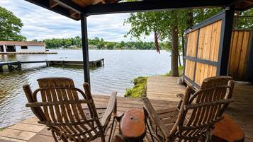 Een houten terras met twee stoelen en een tafel, uitkijkend op een waterpartij met een pier en een huis op de achtergrond.