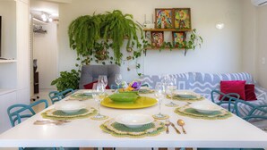 Un comedor con una mesa blanca preparada para seis personas, rodeada de sillas color turquesa. Hay individuales verdes y amarillos, platos blancos y cubiertos. Una planta verde grande cuelga sobre la mesa, y se exhibe arte colorido en la pared.