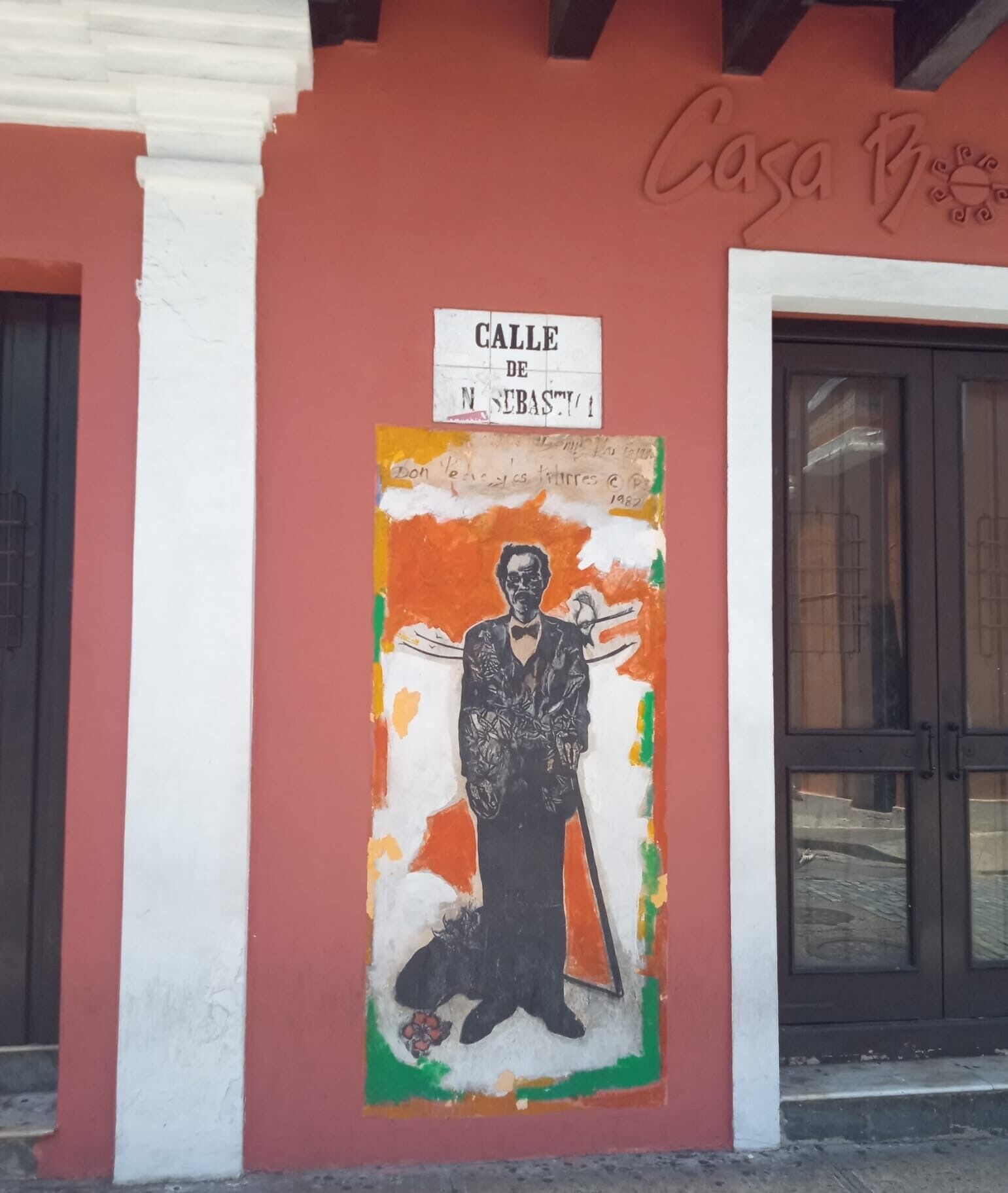 A building facade with a mural of a man in a suit holding a book, a sign reading 'CALLE DE M. NEBASTI,' and a door with glass panels.