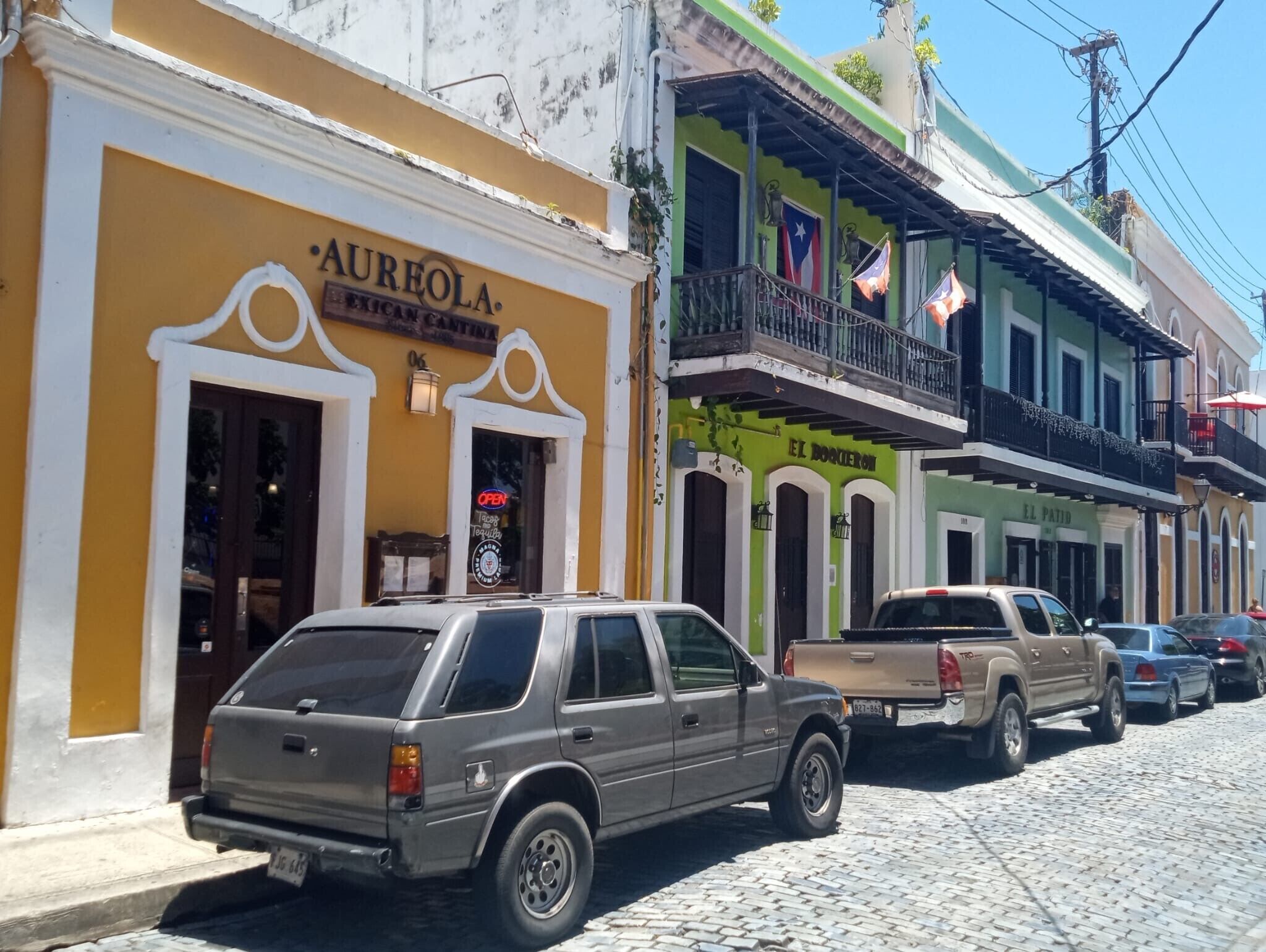 Una calle con edificios, incluyendo un edificio amarillo con letreros 'Aureola' y un edificio verde con letreros 'El Doctor'.