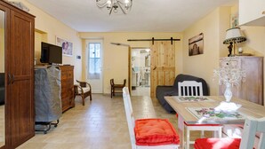 Una habitación con un armario de madera, televisión, una mesa de comedor con sillas, una araña y una cama al fondo.