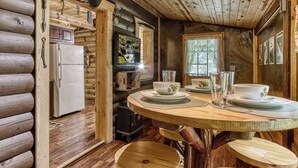 A rustic wooden cabin interior with a wooden table set for dining, a refrigerator, a television, and a window with a view of trees. - Summer Sale! Rustic Romance Classic American Cabin Vibes! (Sevierville)