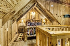 A cozy attic bedroom with wooden walls, a bed with plaid bedding, two bedside lamps, and a wooden railing.