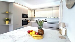 A modern kitchen with white cabinets, a marble countertop, and a fruit bowl with bananas, apples, and a pineapple.