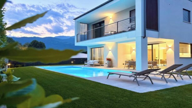A modern two-story house with a swimming pool, outdoor seating, and a view of the mountains.