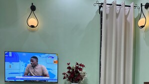A room with a television displaying a news program, a water bottle on a table, a vase with flowers, and a curtain. - Fabulous 1- self contained bedroom apartment with AC in Pwani Region (Kibaha)