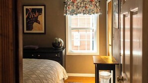Un dormitorio con cama, mesita de noche, ventana con cortina, puerta y una pared con un cuadro enmarcado.