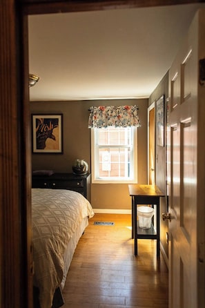 A bedroom with a bed, a nightstand, a window with a curtain, a door, and a wall with a framed picture.