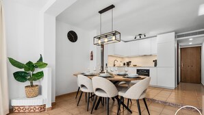 Una cocina moderna con armarios blancos, una zona de comedor con una mesa y sillas de madera, una planta en maceta y un reloj en la pared.