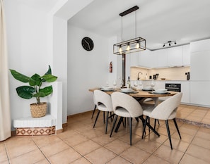 Cozinha moderna com armários brancos, uma área de refeições com mesa e cadeiras de madeira, um vaso de planta e um relógio na parede.