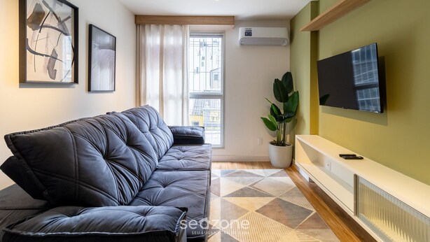 A modern living room with a black leather sofa, a mounted TV, and a large window with curtains.