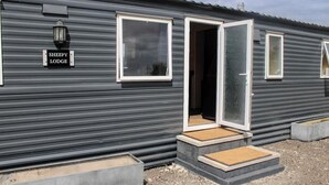 Une maison mobile avec une enseigne indiquant « Sheepy Lodge », dotée de grandes fenêtres, d’une porte et d’un petit espace porche.