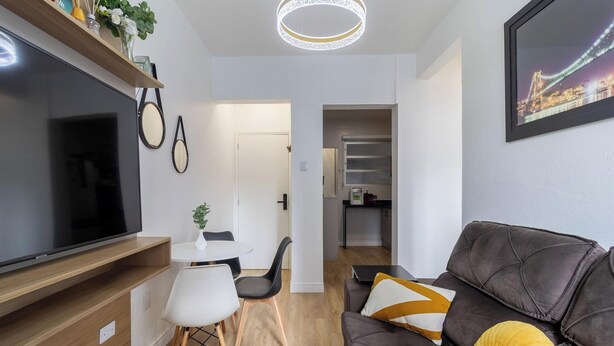 Una sala moderna con televisor de pantalla plana, un sofá con cojines, un comedor con mesa y sillas, y un pasillo que conduce a una cocina.
