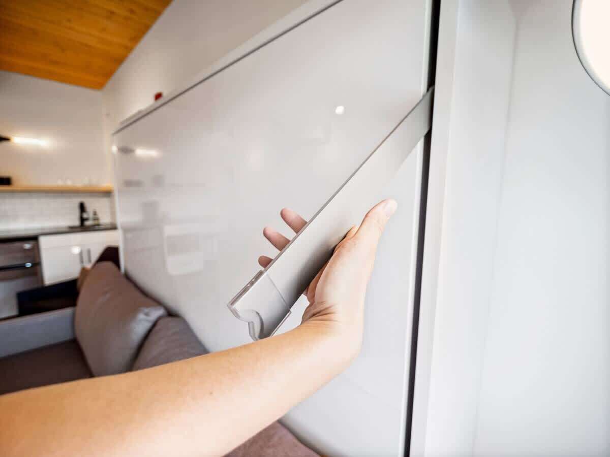 A hand opening a white cabinet door in a modern kitchen with a wooden ceiling.