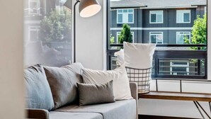 A modern living room with a grey sofa, a floor lamp, and a view of a residential building through a large window. - Uprise 2 Bridgewater Lofts (Spokane)