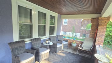 Un patio cubierto con mobiliario de mimbre, una mesita y vistas a un edificio de ladrillo y vegetación.