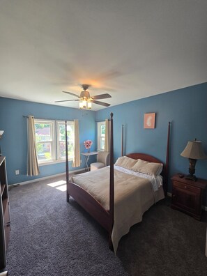 A bedroom with a four-poster bed, a desk, and a chair. There is a ceiling fan, a window with curtains, and a nightstand with a lamp.