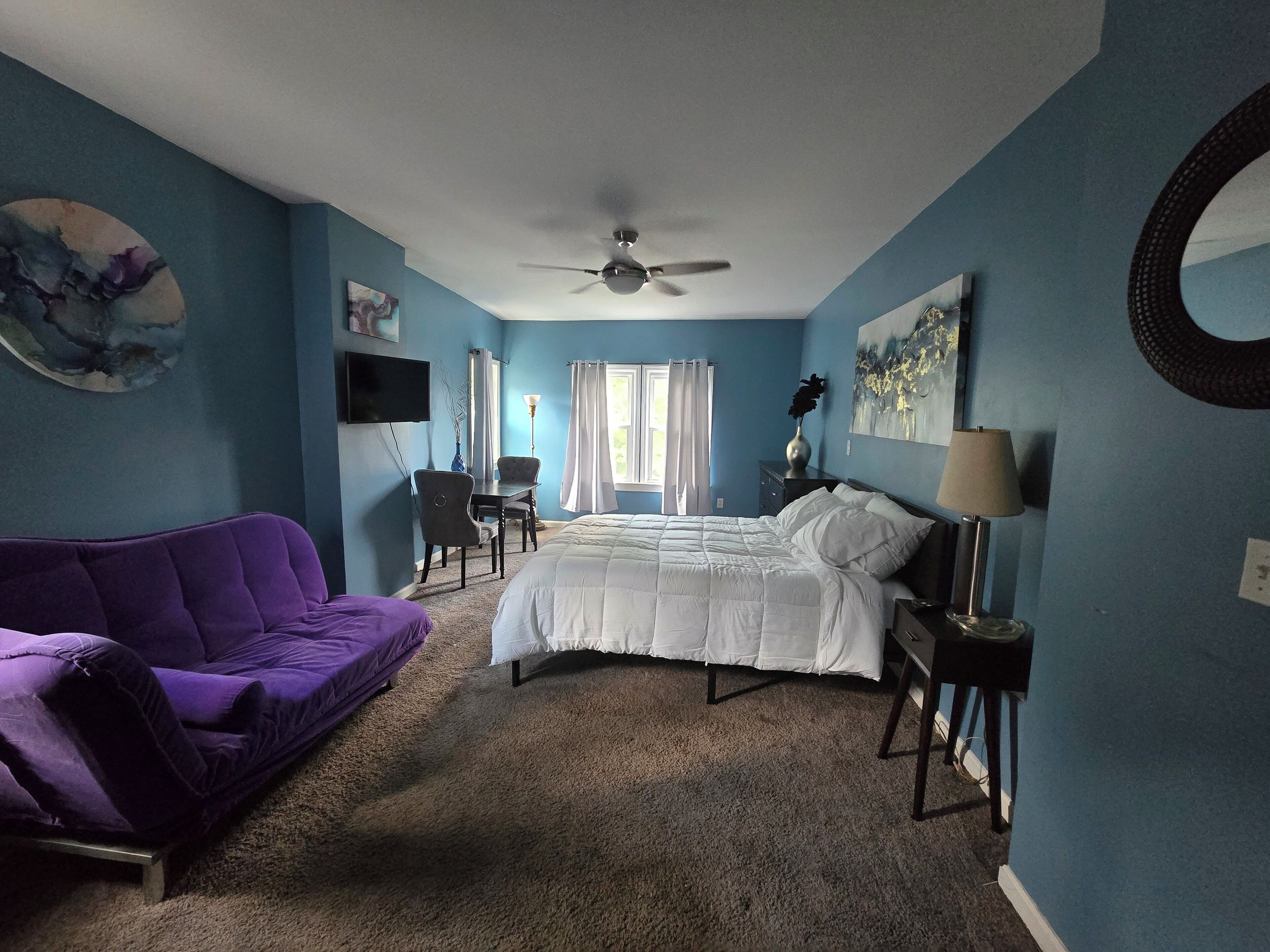 Un dormitorio con cama, un sofá púrpura, un escritorio, una silla, ventilador de techo y obras de arte en las paredes.
