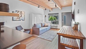 Een moderne woonkamer met een houten plafond, een grijze bank, een houten eettafel en een keuken met een witte koelkast en een koffieapparaat.