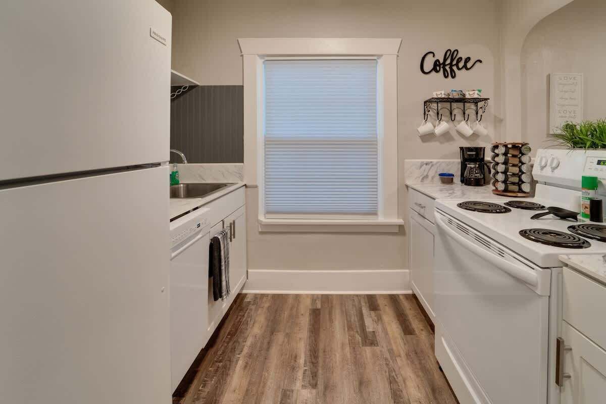 Une cuisine compacte équipée d’un four encastré, d’une plaque de cuisson, d’un espace dédié au café et d’une fenêtre avec des stores.