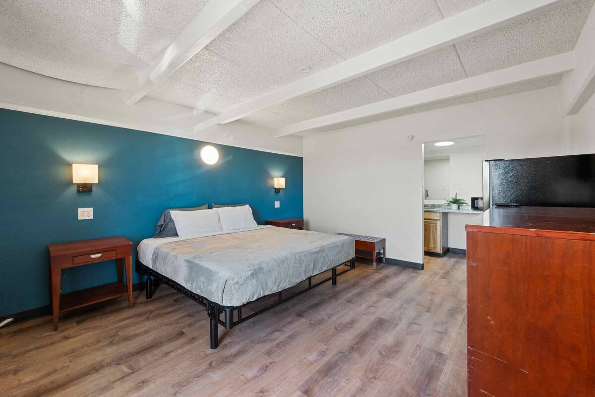 A bedroom with a bed, bedside tables, a nightstand, a desk, a chair, and a kitchenette area.
