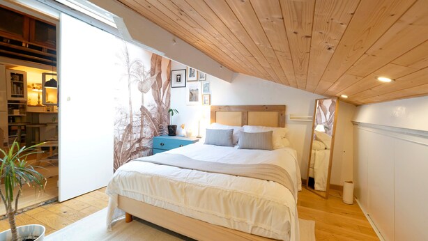 Ein Schlafzimmer mit Holzbalkendecke, einem Bett mit weißer Bettwäsche, einer kleinen Topfpflanze und Blick auf einen Küchenbereich.