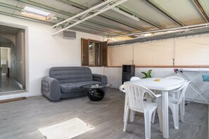 Une pièce avec un canapé gris, une table basse ronde en verre, une table blanche avec des chaises, une fenêtre avec des volets en bois et un haut-parleur.