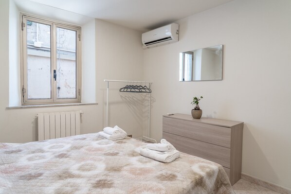 A bedroom with a bed, a radiator, a window, a wall-mounted air conditioner, a dresser, and a mirror.