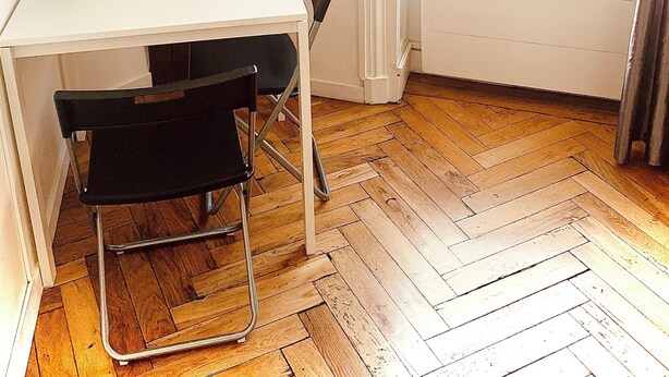 Une petite pièce lumineuse, dotée d’un parquet en bois disposé en point de Hongrie, d’un bureau blanc, d’une chaise noire, d’une machine à café et d’une fenêtre ornée d’une grille en fer décorative.