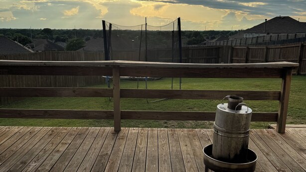 Eine Holzterrasse mit einem Metall-Fassofen, einem weißen Tisch und einem Holzzaun im Hintergrund.