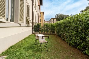 Un pequeño espacio al aire libre con una mesa y sillas, rodeado por un seto cuidado y un edificio con contraventanas.