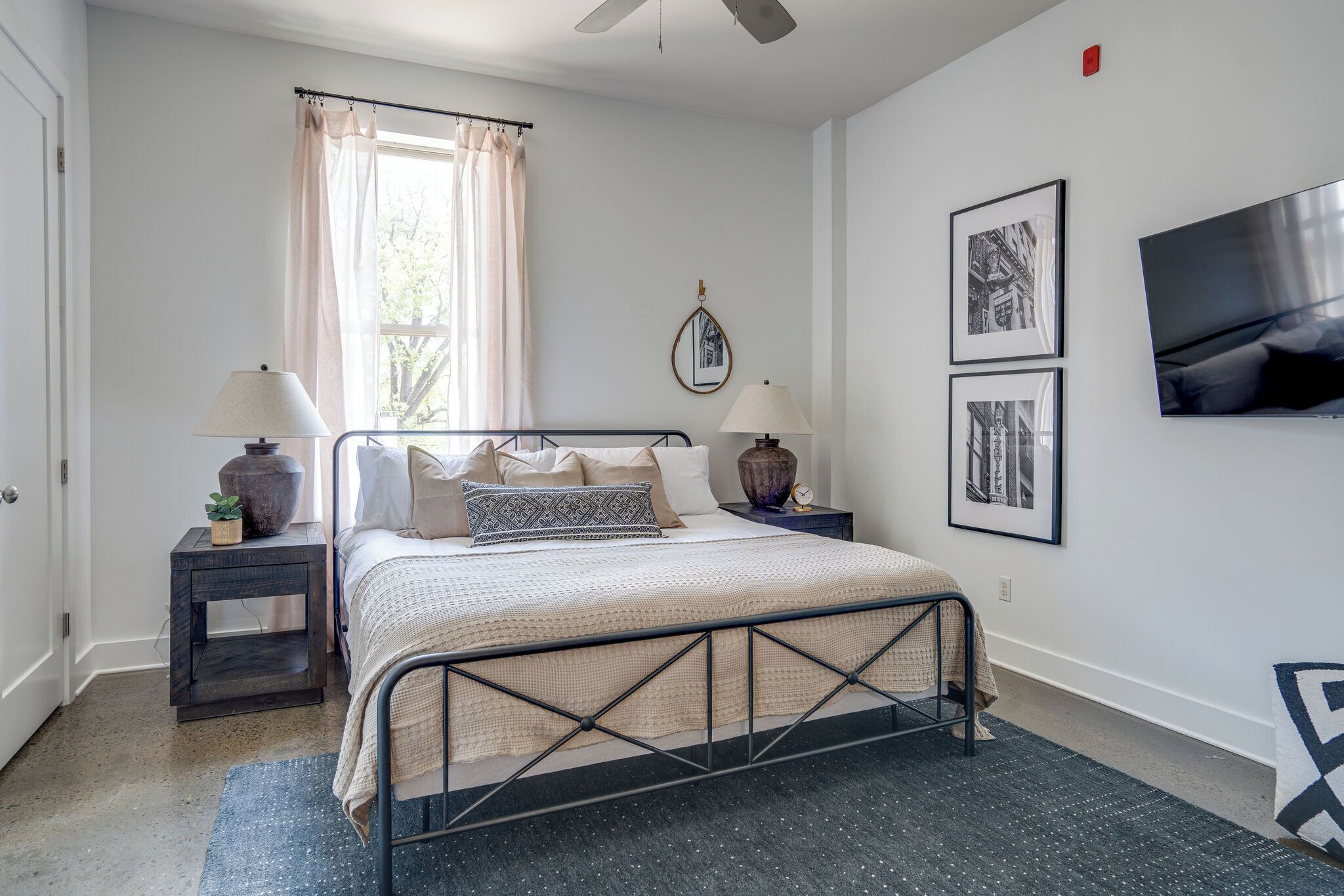 A bedroom with a bed, two bedside tables, a nightstand, a window with curtains, and wall-mounted artwork.