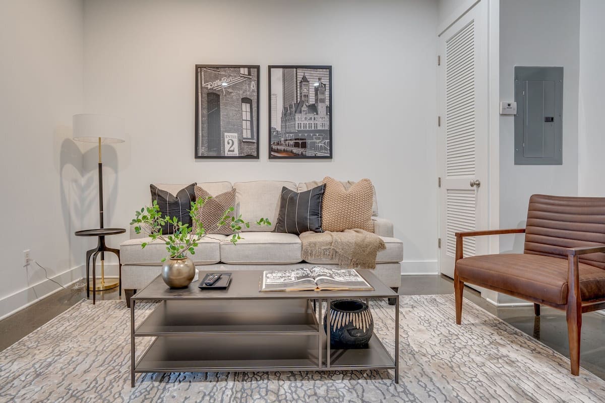 Een moderne woonkamer met een bank, salontafel en een bruine leren fauteuil. Twee ingelijste foto’s hangen aan de muur, een vloerlamp en een klein tafeltje met een plant.