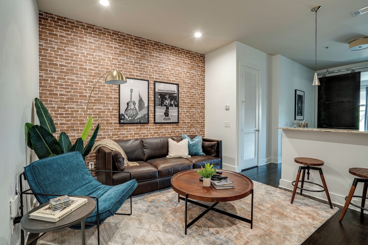 Een moderne woonkamer met een bakstenen muur, een leren bank, een blauwe fauteuil, een ronde salontafel en een grote plant.