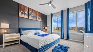 A modern bedroom with a large bed, a nightstand, a dresser, and a view of the city through a large window. - Entertainment Oasis 4BR w/ Home Theater & Game Room (Commerce City)