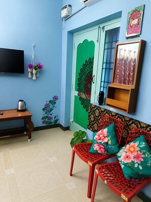 A room with a blue wall, a green door with a peacock design, a red patterned sofa, a wooden table with a kettle, and a TV mounted on the wall. - Blue Waves HomeStay (Puducherry)