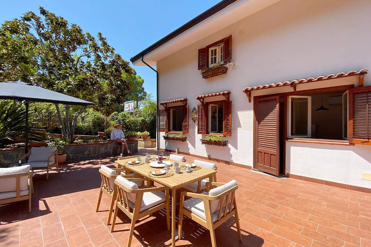 A patio with a wooden table and chairs, a brick floor, and a white building with wooden shutters.