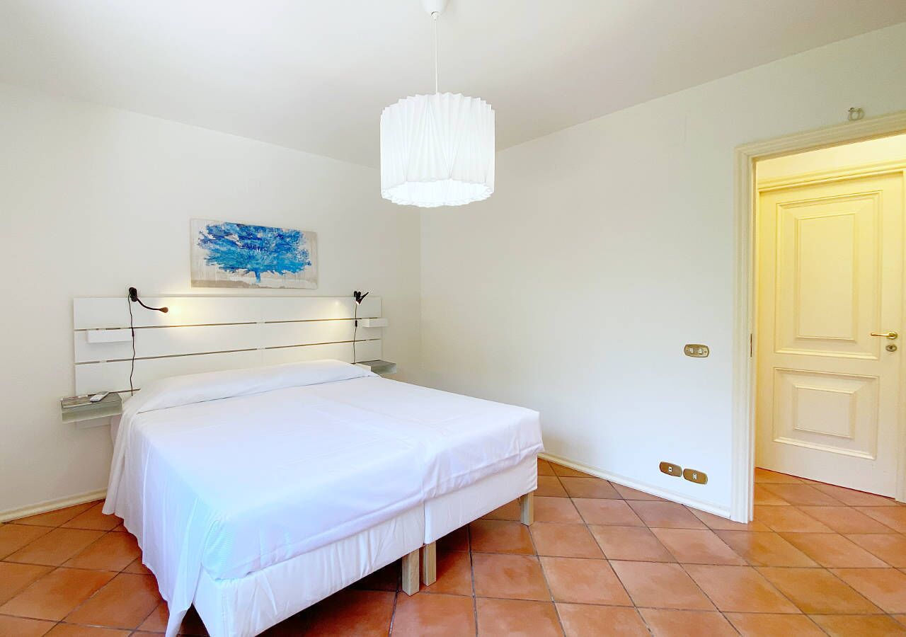 Een slaapkamer met een bed, een witte muur, een schilderij, een witte plafondlamp en een deur.