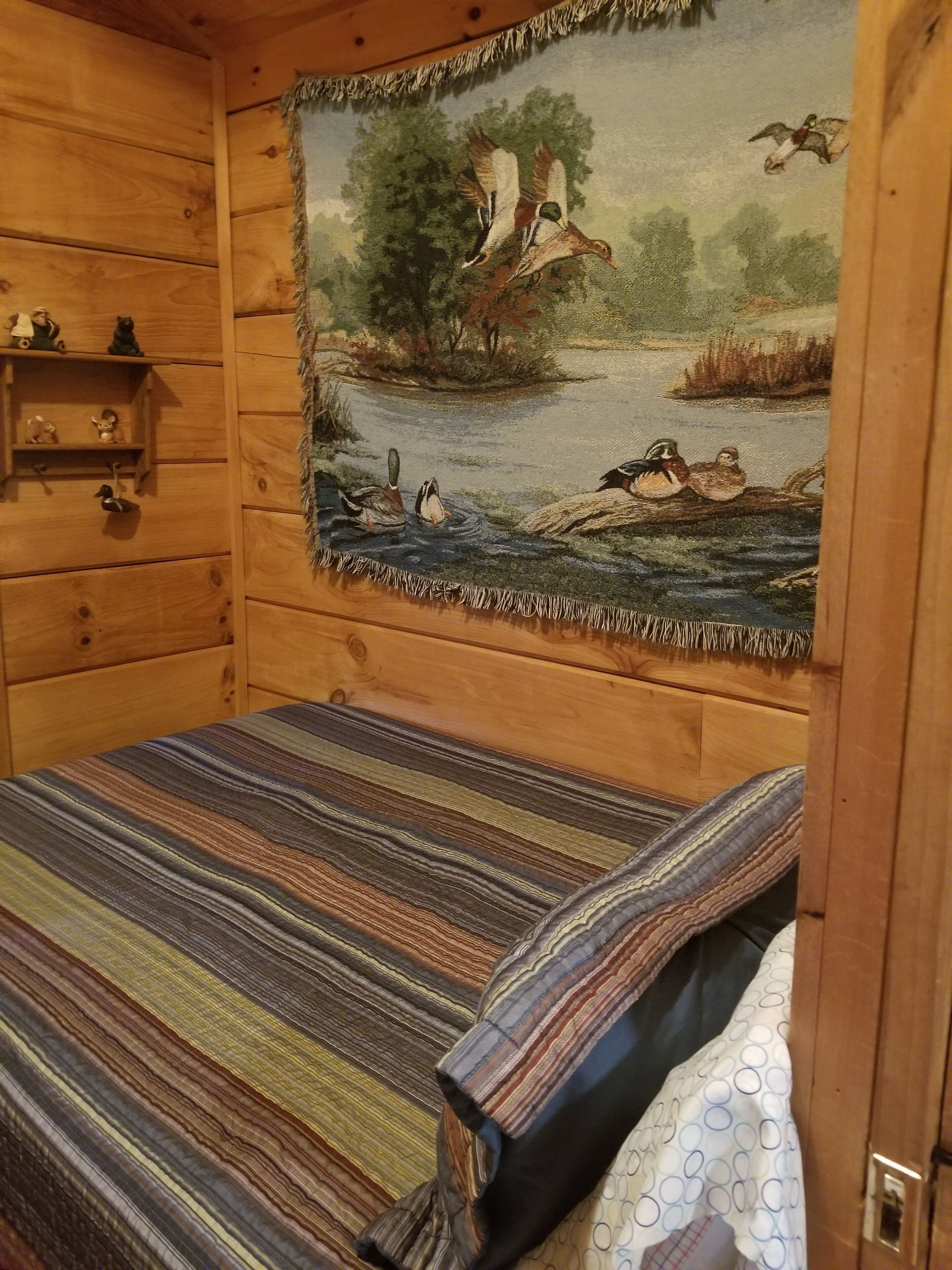A wooden cabin room with a bed, a tapestry depicting a lake scene, and a shelf with small figurines.